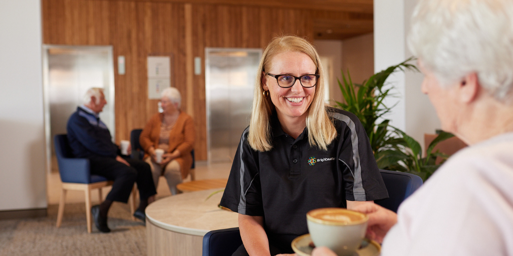 Two women sitting and chatting in cafe area. One woman wears Brightwater shirt.